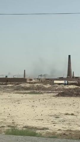 A long shot of a rural industrial area shows several brick buildings with tall smokestacks. Smoke is rising from some of the smokestacks, indicating industrial activity. The ground is dry and dusty, with patches of sparse vegetation. Several power lines are visible overhead. There are a few indistinct figures moving around the buildings, but their actions are not clear. The sky is a pale blue with a few wispy clouds. The overall impression is of a working industrial site in a arid environment.