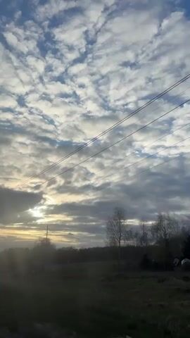 A car is traveling through the countryside at sunrise. Overhead are fluffy altocumulus clouds with glimpses of blue sky. Several utility poles and their accompanying power lines stretch across the frame. Below is a field bordered by a line of trees in the distance. The rising sun shines between the clouds.