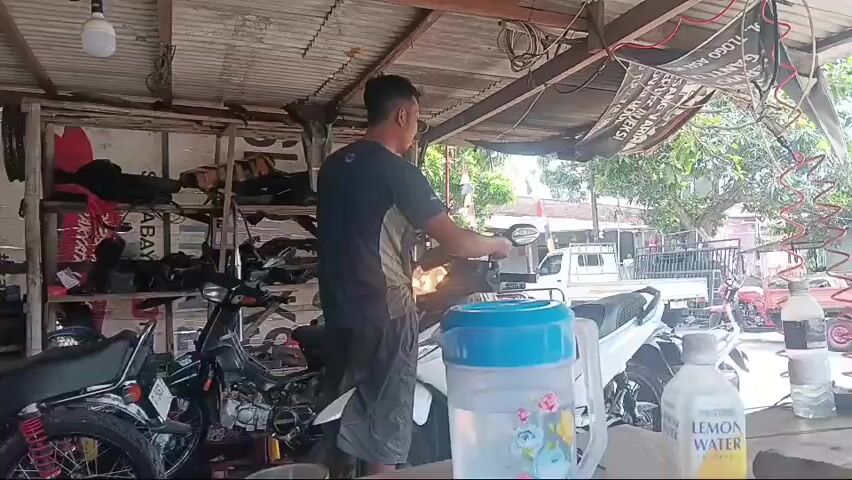 A person is standing in a garage or workshop, holding a small electronic device and looking at a motorcycle. They are wearing a dark t-shirt and light-colored shorts. The motorcycle appears to be a scooter, and it is partially disassembled, with its engine exposed. Shelves filled with motorcycle parts and accessories are visible in the background. A large plastic water jug with a blue lid is on a table in front of the motorcycle, along with a bottle labeled "LEMON WATER." The environment appears to be outdoors, with a corrugated metal roof overhead and trees and buildings visible in the distance. The lighting suggests it is daytime.
