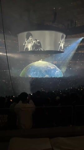 The video shows a large indoor concert venue packed with people. Above the crowd, a giant, illuminated dome resembling the Earth dominates the space. There's a smaller, round screen hanging above the dome, displaying a black and white video of performers. The audience is lit by stage lights and the glow from the dome. Many people in the audience are holding up their phones, presumably recording the performance. A person in the foreground is seated and also appears to be taking a picture. Smoke or haze fills the air, adding to the concert atmosphere.