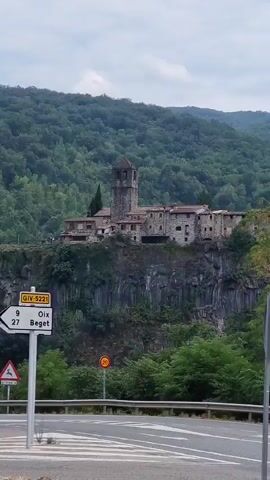 The video shows a view of a historic village perched on a cliff face, surrounded by lush green trees and rolling hills under a cloudy sky. In the foreground, a paved road with lane markings is visible, along with a white traffic signpost and a red octagonal stop sign. A smaller yellow sign with a black symbol is also present. The village itself features stone buildings with terracotta roofs, some of which appear to be very old. A tall, square bell tower rises above the other structures. Trees and vegetation grow around and in front of the village, obscuring some of the lower levels of the cliff. The camera pans slightly to the right, revealing more of the village and the surrounding landscape.