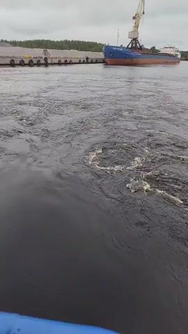 The video shows a body of dark water with visible turbulent currents and eddies. In the background, a blue and white cargo ship is docked at a pier, which is lined with what appears to be a pile of gravel or aggregate. A yellow crane stands on the pier next to the ship. The sky is overcast. The camera appears to be mounted on a moving object, possibly another boat, given the close-up view of the turbulent water in the foreground.