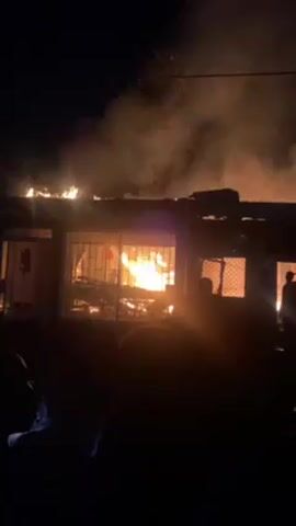 Here's a forensic description of the video:

**Setting:** The video is recorded at night in an outdoor environment. The scene is dominated by a building engulfed in flames.

**Objects:** The primary object is a single-story building, or a section of a building. Visible elements include windows (some with what appear to be security bars), a roof on fire, and general construction materials such as wood or metal frames. Flames are intensely burning, visible both through the windows and on the roof, with plumes of smoke rising above.

**People:** There are multiple individuals present, mostly obscured in the darkness, who appear to be observers or onlookers. Their silhouettes can be seen in the foreground.

**Actions:** The central action is the uncontrolled fire consuming the building. Flames are spreading rapidly, and the structure appears to be significantly damaged. The individuals present are observing and audibly reacting to the fire.

**Environment:** The overall environment is dark, with the primary source of illumination being the fire itself. The air appears smoky, and there may be debris or embers being carried by the wind. The sounds consist of voices, unintelligible conversation, and the crackling/roaring of a large fire.