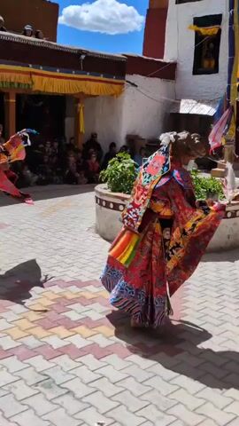 Two individuals are performing a dance in a courtyard. They are wearing elaborate, colorful robes and masks. The person on the right is closer to the camera, while the person on the left is further away and slightly to the left. Both dancers have their arms extended and appear to be moving in unison. The ground is paved with a pattern of pink and yellow tiles. In the background, there is a building with a red and white facade, adorned with yellow and red banners. Several people are seated and standing, observing the dance. The sky is blue with some white clouds.