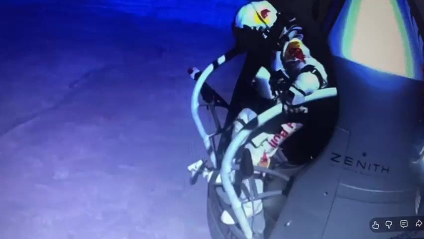 The video shows a person in a white and red spacesuit, branded with "RED BULL", sitting in a capsule with "ZENITH" visible on the side. The person appears to be looking out of the capsule, which is suspended in the air against a dark blue background. The capsule has a metallic frame with curved silver bars. The person's helmet is white with red and yellow accents. The overall impression is that of a space launch or a high-altitude jump.