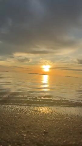 This is a close-up, low-angle video of a calm beach at sunset. The sun is visible on the horizon, casting a golden glow across the water and the sky. The sand in the foreground is a warm, golden color. Gentle waves are lapping at the shore. The clouds in the sky are a mix of dark gray and golden hues, with streaks of light breaking through. There are no people or objects visible in the frame. The overall mood is peaceful and serene.