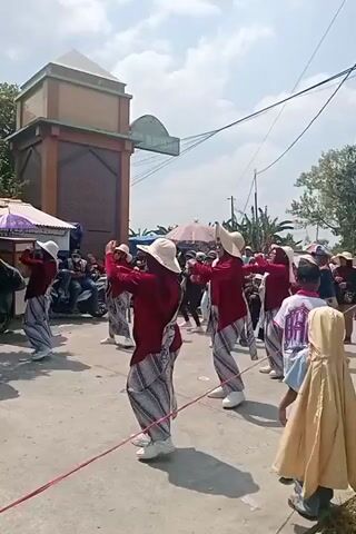 A group of people are performing a synchronized dance on a street. The dancers are wearing matching maroon tops and patterned skirts, with white hats. They are moving in unison, with their arms extended. Several onlookers are gathered on the sides of the street, watching the performance. The scene appears to be taking place outdoors, with a building and trees visible in the background. The sky is clear and sunny. There is a red rope stretched across the street in front of the dancers.