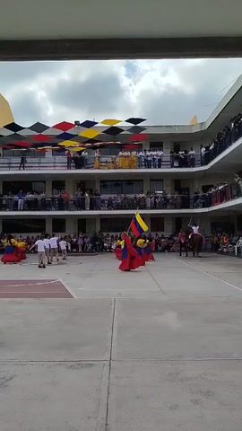 The video captures a vibrant cultural event taking place in a large, open-air courtyard surrounded by multi-story buildings, possibly a school or community center, in Cabudare, Venezuela. The buildings feature balconies from which numerous spectators observe the activity below. Overhead, colorful umbrellas are strung across the courtyard, adding a festive touch.

In the center of the courtyard, several performers are engaged in a traditional dance. They wear elaborate, voluminous skirts in red and yellow, moving gracefully to the accompanying music. One of the dancers carries a flag with prominent yellow, blue, and red horizontal stripes, with the yellow stripe being notably wider at the top, suggesting it could be the flag of Ecuador or Colombia. In the background, another performer is seen riding a horse, further enhancing the cultural display.

A large crowd of people, including adults and children, is gathered on the ground level around the performance area and on the balconies of the surrounding buildings. They are casually dressed, indicating a community event or celebration. The sky overhead is overcast, providing natural, diffused lighting for the daytime event. The overall atmosphere appears festive and engaging.