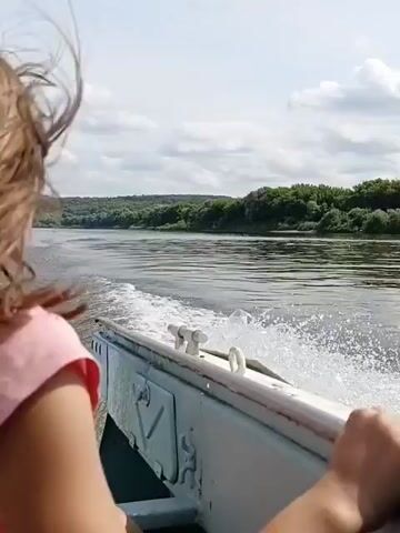 A person's back is visible as they ride in a small boat moving down a river. The person, likely a child, has brown hair blowing in the wind and is wearing a pink shirt. The boat is white and has some metal fixtures on the side. Water is churning behind the boat, creating a wake. The riverbanks are lined with green trees, and the sky is partly cloudy. The camera is positioned behind the person, looking forward along the river.