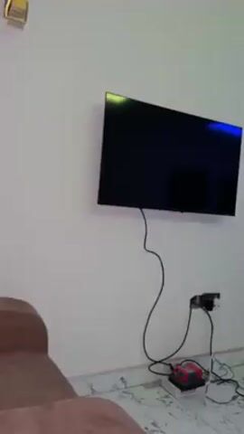 The camera pans upwards from a wall-mounted TV to reveal a living room area. The TV is black and sits above an electrical outlet with some cables and a device plugged in. The room has a brown armchair and a small electric heater. The floor is tiled with a marble-like pattern. There is a set of double doors, and a staircase leading upwards. The ceiling has recessed lighting and a blue light strip. The walls are painted white.