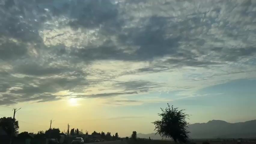The video shows a cloudy sky at sunset with the sun peeking through the clouds on the left side of the frame. In the foreground, there is a silhouette of trees and a landscape. The clouds are a mix of gray and white, with some streaks of blue sky visible. The sun is a bright yellow orb, casting a warm glow on the scene. The overall mood is peaceful and serene.