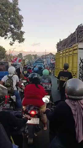 This is a vertical, eye-level, first-person perspective shot of a busy street scene at dusk. The foreground is dominated by the backs of several motorcyclists wearing helmets. The camera is positioned behind them, suggesting the viewer is also on a motorcycle, stopped in traffic. The road is packed with motorbikes, cars, and a large blue truck in the distance. Buildings line the right side of the street, some with signage. Trees are visible on the left, and the sky above displays a warm, hazy sunset with subtle cloud formations. The overall impression is one of a congested urban or semi-urban environment experiencing heavy traffic during the evening commute or a similar event. The lighting suggests it is either sunrise or sunset.