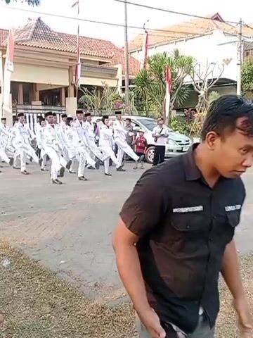 The video shows a group of young people in white uniforms marching in formation on a paved street. They appear to be performing a synchronized drill or parade. The marchers are dressed in white shirts, white pants, and black caps, and they are all moving in the same rhythm. Some of them are carrying what appear to be rifles or batons, but the objects are not clearly visible. The background shows buildings that appear to be houses or other structures, with Indonesian flags displayed. There are also a few cars and a motorcycle parked on the side of the street. The overall scene suggests a formal event or ceremony, possibly a school or military-related activity. The sun is shining, indicating it is daytime, and the sky is clear. The perspective is from the side of the road, slightly behind the marchers.