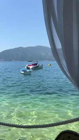 The video shows a scenic view of a bay on a sunny day. The foreground is partially obscured by a white sheer curtain on the right and a white rope railing in the lower part of the frame. The water is clear and turquoise, with sunlight reflecting off the sandy bottom. In the middle ground, two boats are anchored in the bay: a larger white and red motorboat and a smaller white dinghy. In the background, lush green mountains rise from the water, dotted with buildings. The sky is a clear, vibrant blue. The camera pans slightly to the left, revealing more of the coastline and another boat in the distance. The overall impression is one of tranquility and natural beauty.