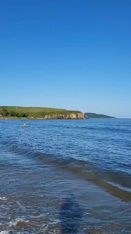 The video shows a serene beach scene with clear blue skies and calm blue water. Gentle waves lap the shore. In the distance, a rocky, green-topped cliff juts out into the sea. Several people are visible swimming and enjoying the water. One person is standing waist-deep in the water, while others are further out, some appearing to be swimming. The water is clear enough to see the bottom in some shallow areas, with some dark seaweed visible. The overall impression is one of a peaceful and pleasant day at the beach.