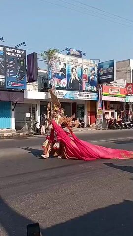A young woman dressed in an elaborate, traditional costume with a large feathered headdress and a long, flowing train walks down a street. She is smiling and posing for the camera. Several people are watching her, and some are taking pictures. The scene takes place on a sunny day, with buildings and advertisements visible in the background. A few motorcycles are parked on the side of the road.