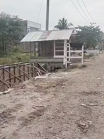 The video presents a daytime view of a roadside scene in Barokulamgajah, Indonesia, under an overcast or hazy sky. In the foreground, there is a dirt path or unpaved road. To the left, a wooden structure, possibly a retaining wall or part of an embankment, runs alongside a ditch or small canal.

Dominating the midground is a small, simple building or hut, constructed on stilts above the water. It features a corrugated metal roof and white-painted wooden railings or framing. Behind the hut, a tall utility pole with multiple overhead wires stands prominently. In the background, a paved road is visible with several vehicles, including cars and possibly a bus, driving past. Trees and other distant buildings complete the rural or semi-urban landscape. There is no discernible activity or visible text in the scene.