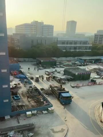 The video shows a construction site with a group of workers unloading a truck filled with bamboo scaffolding. The workers are wearing hard hats and are using hand carts to move the bamboo. The site is located next to several buildings, and there is a crane visible in the background. The weather appears to be sunny.