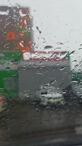 The video shows rain droplets clinging to a window, obscuring the view of the outside. Through the water-streaked glass, a blurred gas station is visible, with green and red colors prominent. The scene suggests a rainy day, likely viewed from inside a vehicle due to the presence of the window and implied movement. Overall, the setting is somewhat dreary and indistinct because of the rain and focus on the wet window.
