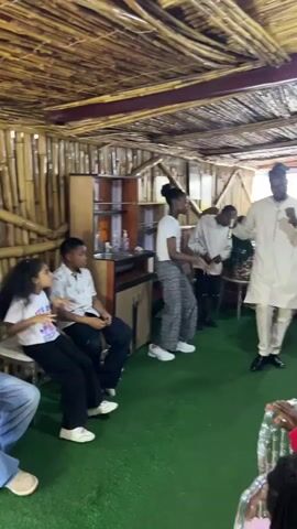 A group of people are in a room with a bamboo ceiling and walls. In the foreground, a young girl with curly hair is sitting on a chair, facing left. To her right, a young boy in a white shirt and black pants is also sitting. Further back, several children are standing and appear to be dancing or moving to music. A man in a white traditional outfit is standing on the right side of the room, facing the children and gesturing with his hands. The floor is covered with green carpet. There is a cabinet with shelves in the background, and some chairs are visible on the far right.