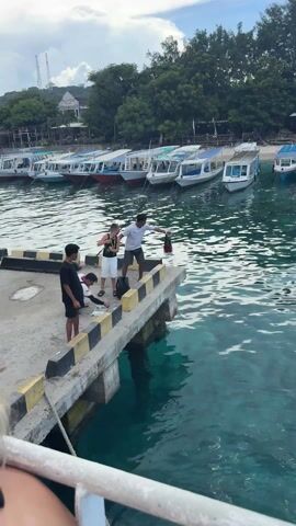 A medium shot shows four people on a concrete pier.  One boy, wearing a black tank top and white shorts, holds a fishing pole and reaches for a backpack on the ground. An adult man, wearing a white t-shirt and gray shorts, stands next to him, holding a red-and-black striped bag. To the boy’s left, one person in a black t-shirt stands watching, while another person in a red-and-white striped shirt leans over the ground, apparently preparing fishing tackle. In the background are several small boats tied to the pier, and across the water are trees and buildings. The water is turquoise and rippled. The day is overcast.