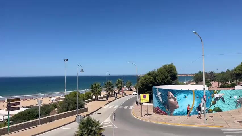 The video opens with a wide shot of a beach on a sunny day. The ocean is a deep blue with moderate waves. A sandy beach is populated with numerous beachgoers, lounge chairs, and umbrellas. A road curves along the coastline, with palm trees and vegetation on either side. A large, colorful mural depicting a woman's face and birds is visible on a circular structure next to the road. Several streetlights are positioned along the road. Cars and people are seen walking and driving on the road. The sky is a clear, bright blue. As the video progresses, the camera pans slightly to the left, offering a more expansive view of the beach and ocean. The road continues to curve, with more palm trees and greenery. People are seen walking on the sidewalk adjacent to the road. A stop sign is visible. The overall scene depicts a lively and scenic coastal environment.