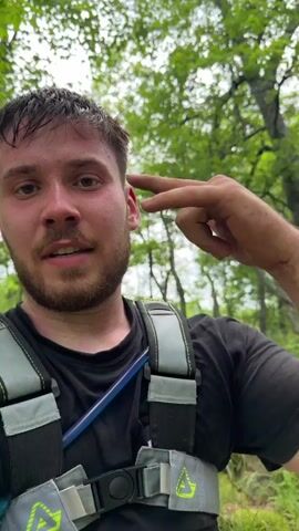 A man in a black shirt and gray chest harness takes a selfie, giving a peace sign. A second man in a blue shirt and black backpack rides a dirt bike through a forest trail. The man on the dirt bike, wearing a helmet, navigates over rocks and through dense green foliage. He briefly stops the bike before continuing to ride downhill.