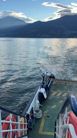 A ferry is moving forward on a body of water. The camera is positioned on the deck of the ferry, looking backward towards the stern. A wake trails behind the ferry, creating frothy white patterns in the water. In the distance, mountains covered in vegetation rise from the opposite shore. The sky above is a pale blue with scattered clouds, some of which are illuminated by sunlight. On the ferry deck, there are safety markings, life preservers, and coiled ropes. The ferry appears to be traveling across a lake or a large river. The scene is captured during what appears to be either early morning or late afternoon, given the angle of the sunlight and the muted colors. No people are clearly visible in the frame.