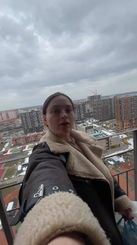 A woman with brown hair pulled back, wearing glasses and a brown jacket with a cream-colored shearling collar, takes a selfie on a balcony. She makes an exaggerated surprised face, then puckers her lips. The background features a cityscape with modern apartment buildings, a school with colorful playgrounds, and patches of snow on the ground. The sky is overcast. The woman is holding a phone in her right hand to record the selfie and a white plastic bag in her left. The balcony has a metal railing.