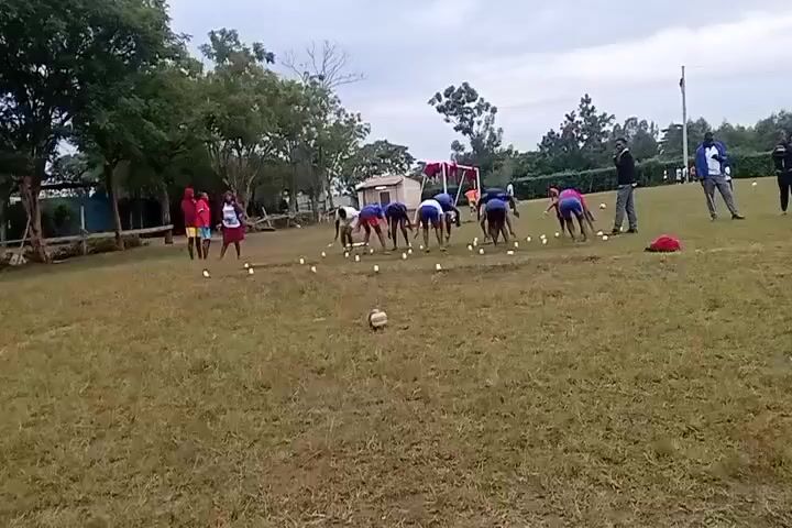 A group of people are engaged in a physical activity on a grassy field. There are about a dozen people bent over, placing what appear to be small, white objects, possibly eggs or balls, on the ground in a line. Several other individuals are standing around observing, including a few adults who might be coaches or instructors. The people bending over are wearing athletic clothing, and the people standing appear to be wearing everyday clothing. There is a soccer ball sitting in the grass. Behind the group, trees, a small building, and a playground structure are visible. The sky is overcast.
