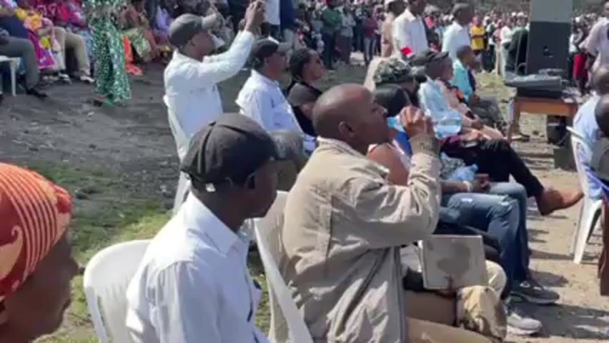 Here is a concise description of the video:

The video shows a crowd of people sitting on chairs outdoors, likely attending an event. Most people are looking in the same direction, presumably towards a speaker or stage. Some individuals are taking pictures with their phones. One person is drinking from a water bottle. A speaker sits on a table near the crowd. The environment appears to be somewhat grassy or unpaved, and there is a building visible in the background. The overall atmosphere seems to be one of anticipation and attentiveness.