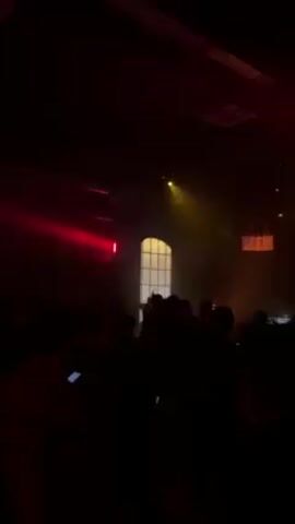 The scene is a dimly lit, large interior space filled with a crowd of silhouetted people. A single figure is visible in front of them, illuminated by spotlights; this person appears to be a DJ or performer standing behind a sound mixing table. The venue has high ceilings and a tall, arched window toward the back. Decorative structures covered in fabric hang from the ceiling, and lighting effects in shades of red and yellow create a dynamic atmosphere. The audience is predominantly dark, but some individuals are holding up phones, presumably to record or photograph the performance. Overall, the environment suggests a concert or a club with a live music performance.