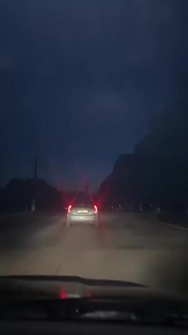The video shows a view from inside a car at dusk or night. The car is driving on a road, following another car. The taillights of the car ahead are red and visible. The sky is dark blue and cloudy, with some trees lining the sides of the road. The headlights of the car are on, illuminating the road ahead. There are some smudges or water droplets on the windshield of the car from which the video is recorded.