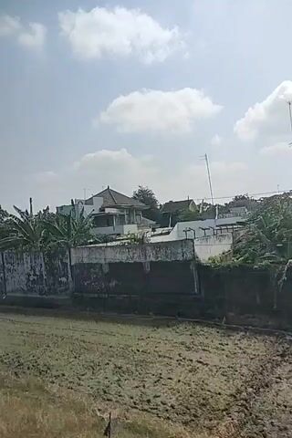The video shows a view from a moving vehicle, looking outwards towards a landscape under a bright, cloudy sky. In the foreground, there is dry, light-colored grass or weeds. A low, weathered concrete wall runs horizontally across the middle of the frame, behind which are trees and some buildings. The buildings appear to be simple, concrete structures, some with visible windows. One building is a two-story structure with a metal roof. The wall itself is discolored and shows signs of age and weathering. The trees are green and leafy, with some appearing to be banana trees. The overall impression is of a somewhat rural or industrial setting.