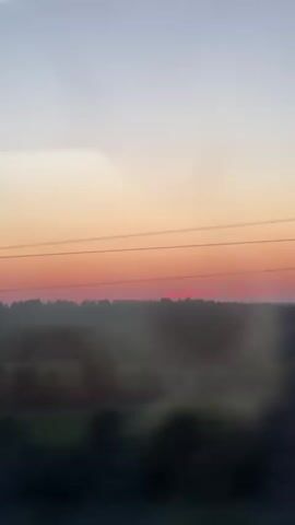 The video shows a blurry, fast-moving view of a landscape at sunset. The sky transitions from pale blue to orange and pink near the horizon. A dark line of trees is visible in the distance. The foreground is obscured by dark, out-of-focus shapes, possibly trees or foliage passing by. The motion suggests the video was recorded from a moving vehicle. There are no identifiable people or specific objects. The overall impression is one of a fleeting, abstract glimpse of nature.