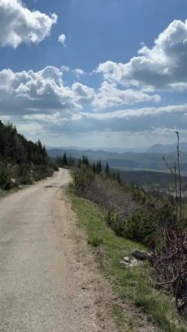 The video shows a scenic mountain landscape under a partly cloudy blue sky. The camera pans across a view of rolling hills and distant mountains, partially covered in forests. A dirt road is visible in the foreground, lined with vegetation. There are no people visible in the video.