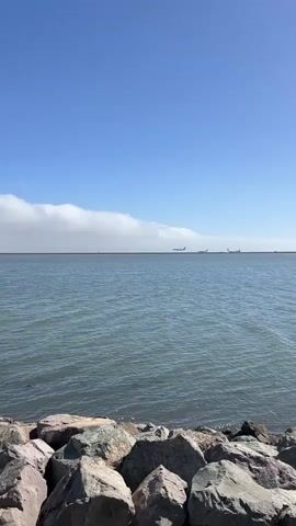 A video taken from a rocky shoreline looks out over a large body of water. The sky is clear blue, with a few wispy clouds stretching across the horizon. Two airplanes are visible in the distance, flying over the water. The waves are calm, and the water appears to be dark blue. The rocks in the foreground are varied in size and color, with some appearing to be gray and others reddish-brown.
