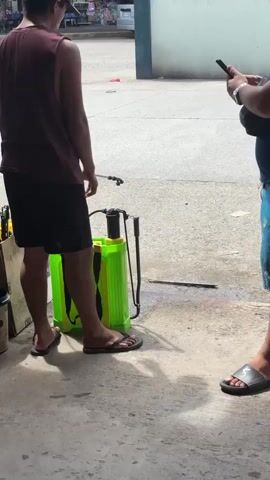 A man wearing a maroon tank top and black shorts stands with his back to the camera. He is wearing black flip-flops. He holds a sprayer in his right hand, connected to a green backpack sprayer. To his left is a collection of buckets, one of which is yellow. To his right, another person is partially visible, wearing a black shirt and blue shorts, holding a black smartphone. They are standing on a concrete surface, with shadows suggesting an outdoor, sunny environment.