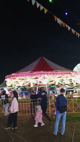 At night, an amusement park is open. A carousel with a red and white striped canopy, spinning fast and lit with bright lights, is visible, surrounded by a yellow barricade. Several people are standing around watching the ride, including a person in a black jacket, one in a pink outfit, and another in blue jeans. Bunting hangs overhead in the dark sky. Another lit-up ride is visible in the background.