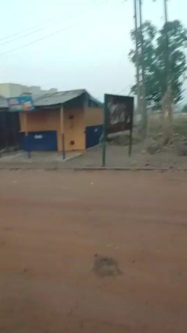 The video shows a dirt road lined with small, run-down kiosks or stalls. The kiosks are made of wood and metal sheeting, and some have signage on them. A street lamp stands next to one of the kiosks. The road is dusty and littered with debris. In the background, a person can be seen walking in the distance. The sky is overcast and the overall impression is one of a poor or underdeveloped area. The camera pans slightly to the left as the video progresses.