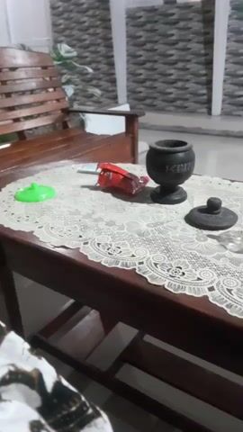 The video shows a close-up, medium shot of a wooden table with a lace tablecloth. On the table, there is a red package, a green lid, a small black urn with a lid, and a clear glass mug with some brown liquid and ice. The background shows a wooden bench, a potted plant, and a textured wall. The lighting suggests it is daytime or evening with artificial light. The camera angle is slightly from above, looking down at the table.