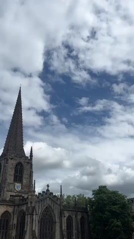 The video shows a tall, gray stone church spire against a sky filled with a mix of blue and white clouds. The church is partially visible, showcasing its Gothic architectural style with arched windows and intricate details. A clock is visible on the spire. A tree with green foliage stands to the right of the church. The camera angle is looking up at the church, emphasizing its height and grandeur.