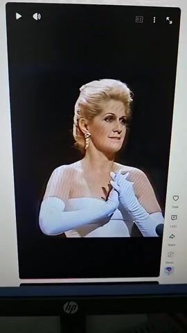 A woman in a white strapless gown and long white gloves stands on a stage with her hands clasped in front of her. A man in a suit sits at a grand piano to her left. The woman looks to her right and smiles. She then touches her chest. The man at the piano turns and looks at the woman. The woman turns her head and looks at the man. She then looks forward again and begins to sing. A man in the audience with glasses and a bald head wipes his eye.