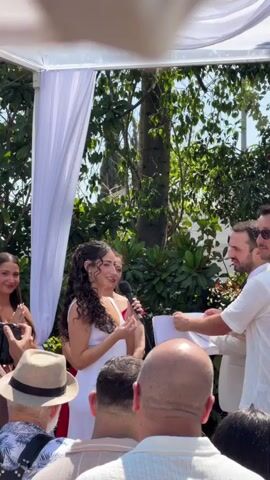 A wedding ceremony is taking place outdoors under a white canopy. A woman with long, curly dark hair, wearing a white dress, stands holding a microphone and speaking. To her right, a man in a light-colored suit stands facing her, holding a piece of paper. They are exchanging rings. In the background, lush green foliage and trees are visible. Several people are watching the ceremony, some with cameras. One person in the foreground is wearing a hat. The lighting suggests it is daytime.