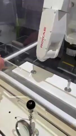 A person's hand is holding a thin, white rod near a white robotic arm labeled “ROKAE” inside a glass enclosure. The robotic arm is positioned above a white surface with three raised silver pegs and a thin, white line running between two of the pegs. In the foreground, a magnifying glass on a stand is visible. The person manipulates the rod, seemingly interacting with the area under the robotic arm's precision point. The environment appears to be a public space or exhibit, with other people visible in the blurred background.