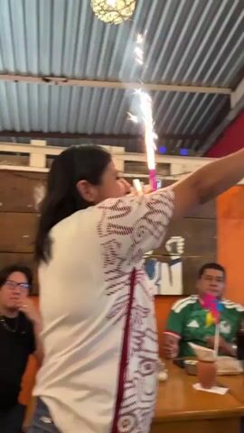 The video opens with a woman in a white and red patterned shirt holding a drink with a sparkler in it. She is looking forward and appears to be celebrating. The background shows a ceiling with textured panels and lighting fixtures. The scene then pans to the right, showing more people in a festive environment. There are colorful banners, flags, and decorations visible, including what appears to be a television screen in the background. Several people are wearing green shirts, suggesting a sporting event, likely a soccer match, given the presence of a soccer ball decoration and flag colors that match national teams. The camera continues to move, showing a man smiling and looking at the camera, then pans to show a group of people at tables, some cheering with their hands raised. The overall atmosphere is lively and celebratory.