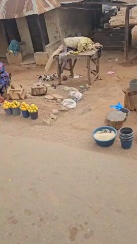The video shows a market scene in a developing country, likely Africa, from the perspective of a moving vehicle. Several vendors are seated or standing next to their goods, which include limes, pineapples, yams, and mangoes, displayed in buckets and piles. Buildings with corrugated metal roofs line the dusty roadside. People, including children, are moving about the market area. The vehicle honks as it passes by.