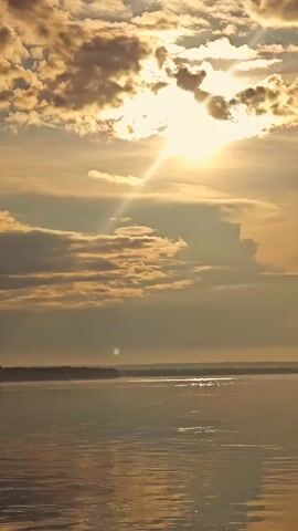 This media file features a serene outdoor scene at sunset. The sky is filled with a dramatic display of clouds, illuminated by the golden rays of the setting sun. Shafts of light pierce through the clouds, creating a warm and ethereal glow. Below the sky, a vast body of water, likely a lake or a wide river, reflects the colors of the sunset. The water's surface is calm with gentle ripples. In the distance, a faint outline of a treeline is visible along the horizon. A small, dark object, possibly a buoy or a small boat, can be seen on the water to the left of the frame. The overall mood is peaceful and awe-inspiring, evoking a sense of tranquility. The time of day is clearly sunset, indicated by the low sun and the warm, golden light. There is no visible text in the media file.
