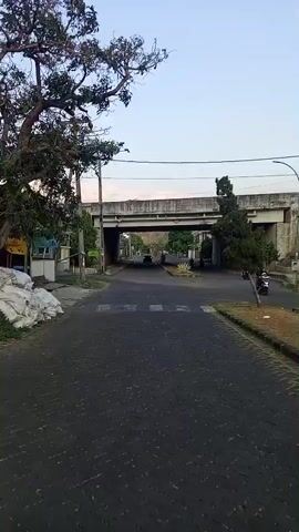A camera moves forward along a paved road, which is covered in dark cobblestones. The road is bordered on the left by a sidewalk and a small building with a blue awning, and on the right by a grassy area with a few trees and a motorcycle. A traffic light is visible on the left side of the road. In the distance, the road goes under a concrete overpass. A dark-colored car is driving under the overpass. Two people on a motorcycle are also visible near the overpass, to the right of the road. The sky is light blue and overcast.