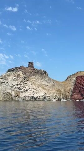 This video captures a picturesque view of a rocky island coastline from the water. A weathered, ancient tower stands prominently atop a rugged, pale rock formation. To the right of this main rocky outcrop, a steep cliff face displays striking red-hued rock layers. The clear blue sky is dotted with wispy white clouds, suggesting fair weather. Several white sailboats are anchored in the calm, deep blue water, their masts adding a touch of elegance to the natural scenery. The sun is high, indicating it is likely midday. The overall impression is one of natural beauty, tranquility, and historical significance. The location appears to be Capraia Isola, Italy, a volcanic island known for its dramatic cliffs and clear waters, offering a serene escape.